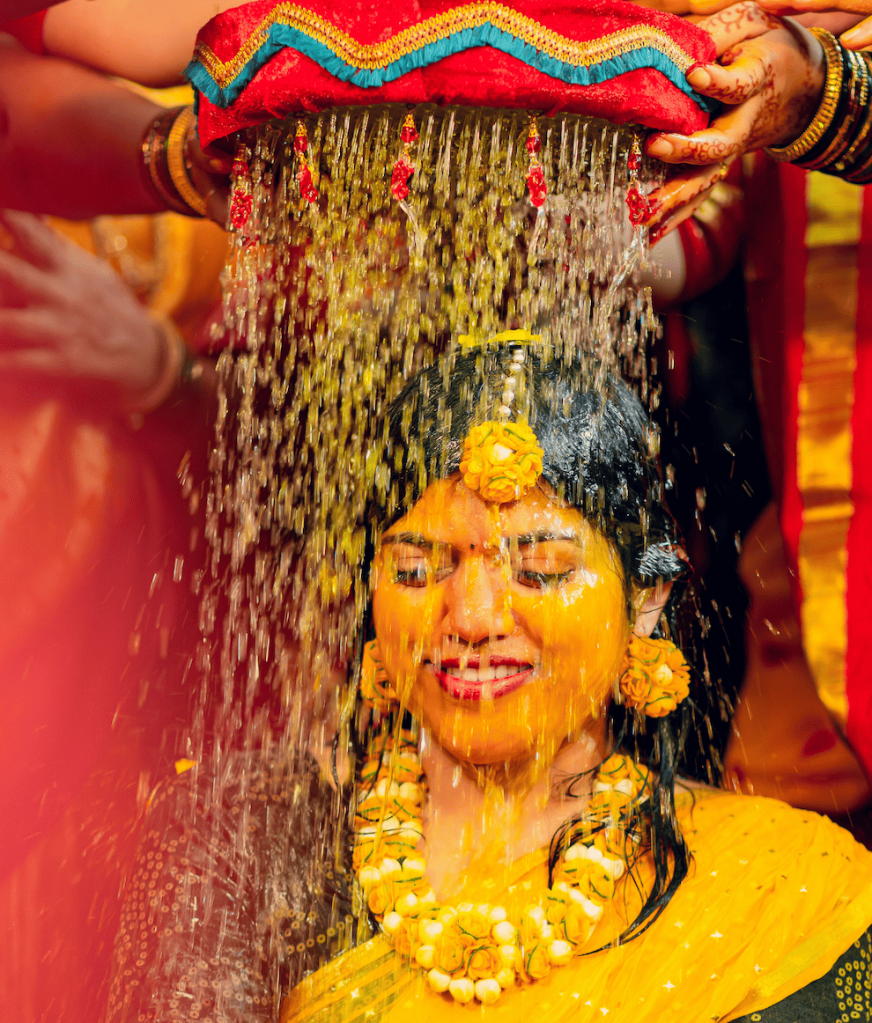 Une femme souriante portant un masque capillaire au curcuma lors d'une cérémonie traditionnelle, entourée de décorations colorées et de symboles culturels.
